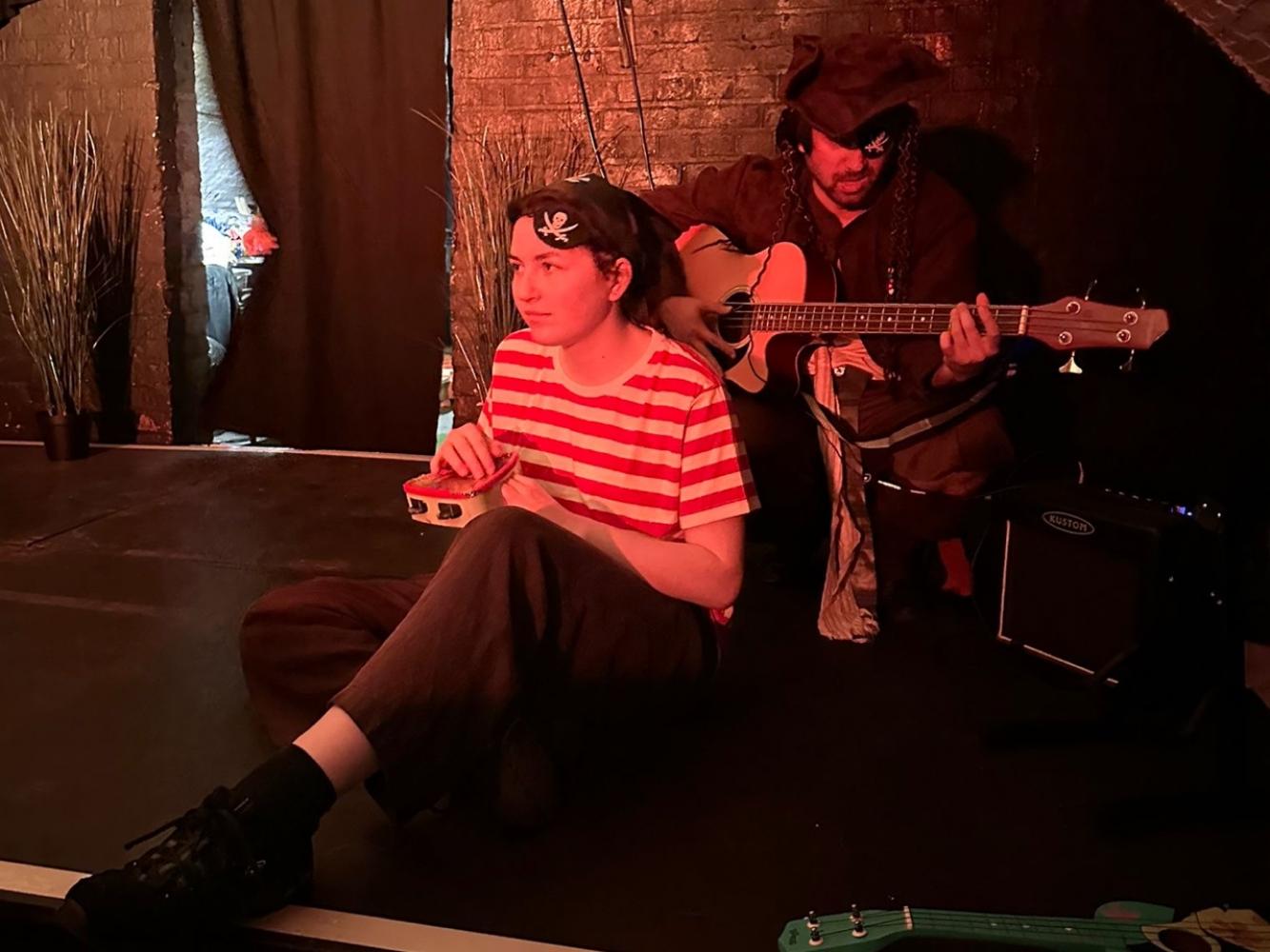 2 pirates sit on the floor, one is playing the base guitar, while the one in front is playing a tamborine