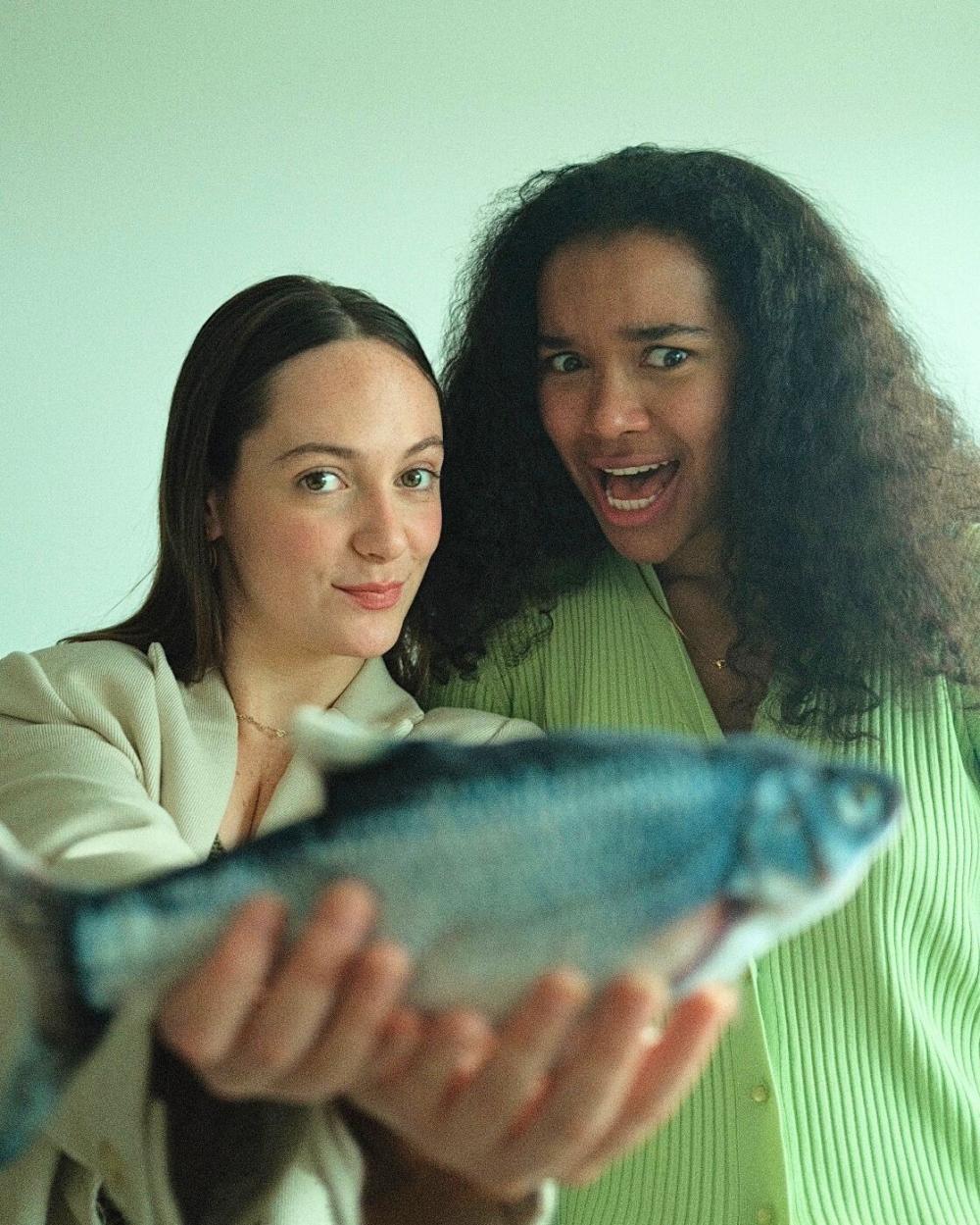 Two women stand side by side. Woman on the left holds a fish to camera and looks smug. Woman on the right looks bewildered.
