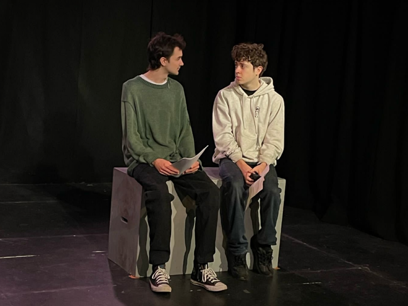 2 men sit on some grey cubes.  They are looking at each other, there is an awkward energy about them both.