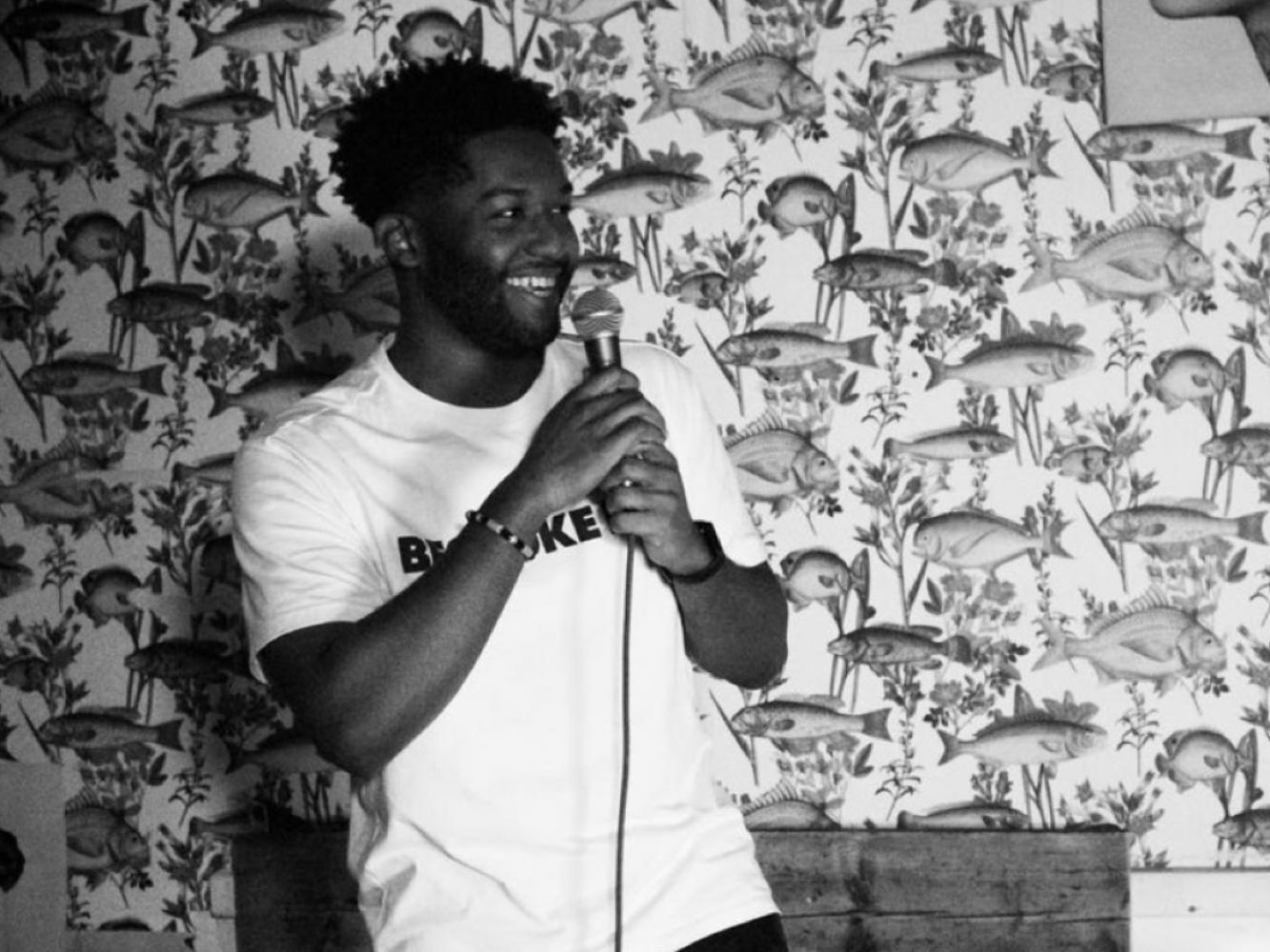 A man stands against a brick wall hoping a microphone, he is smiling and the cable runs down the centre.