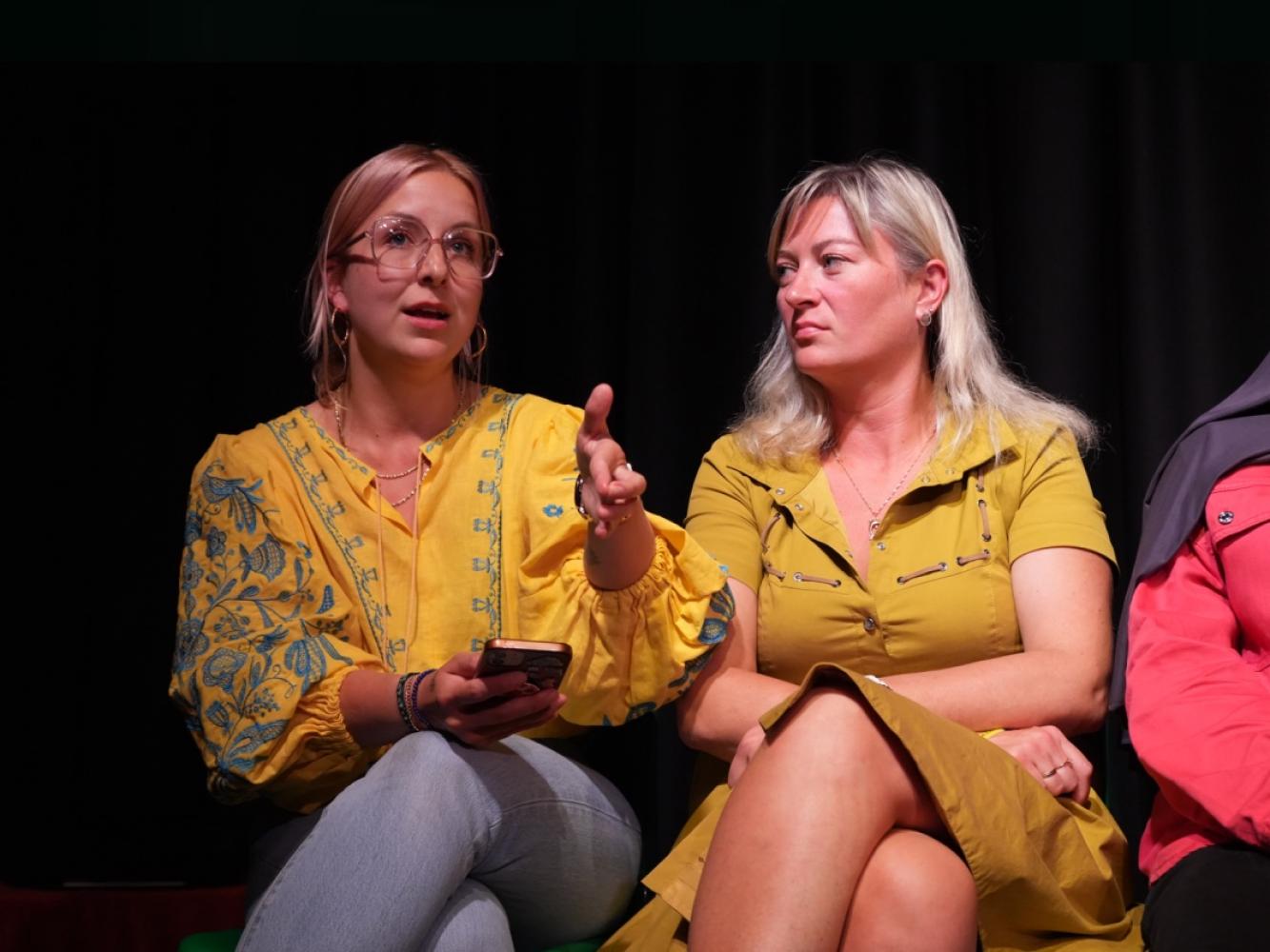 Two women sit in conversation.  One has a phone in her hand the other listens to her.  They are both in Yellow.