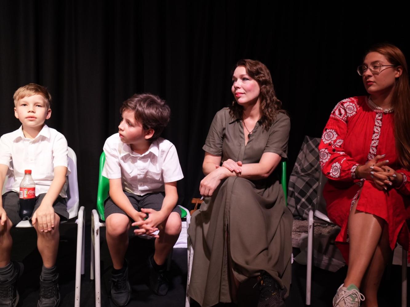 4 people sit in a row, 2 children dress in white and 2 women dressed in traditional dress.  They are all in conversation.