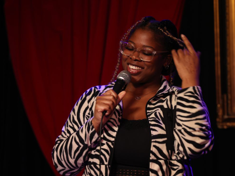 Woman in glasses smiling with a microphone, touching her hair, dark red curtains in the background