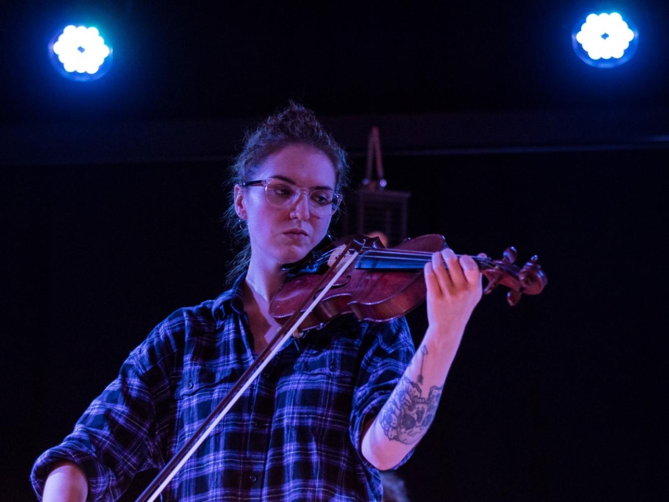 A Violin player stands playing her instrument.  She is lit with blue light and wearing a blue check shirt.