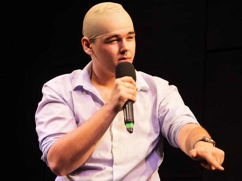 Drag King doing stand-up comedy to the crowd about business and making money.