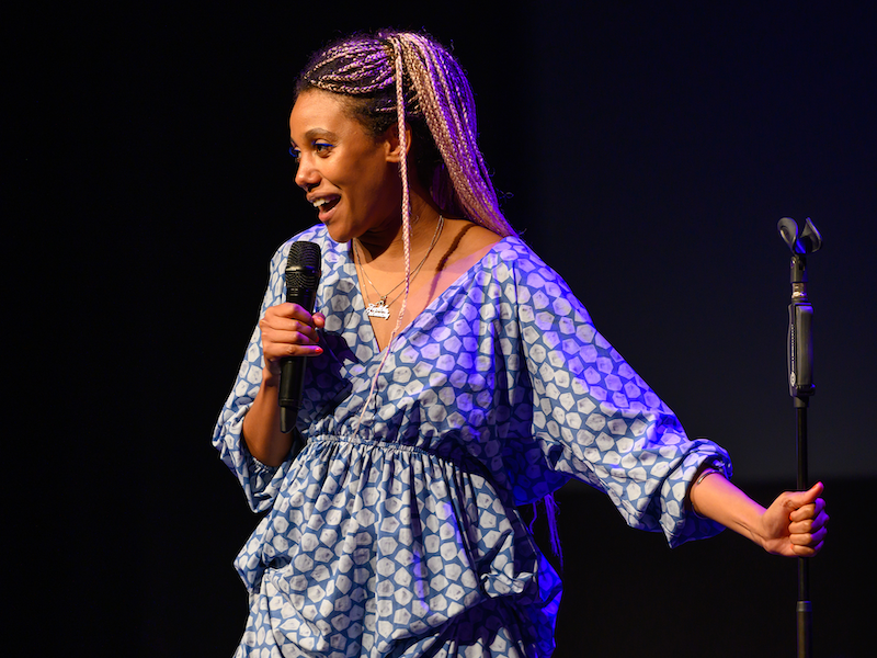 Kate Cheka in a blue-patterned dress and pink braids holding a mic in one hand and a mic stand in the other