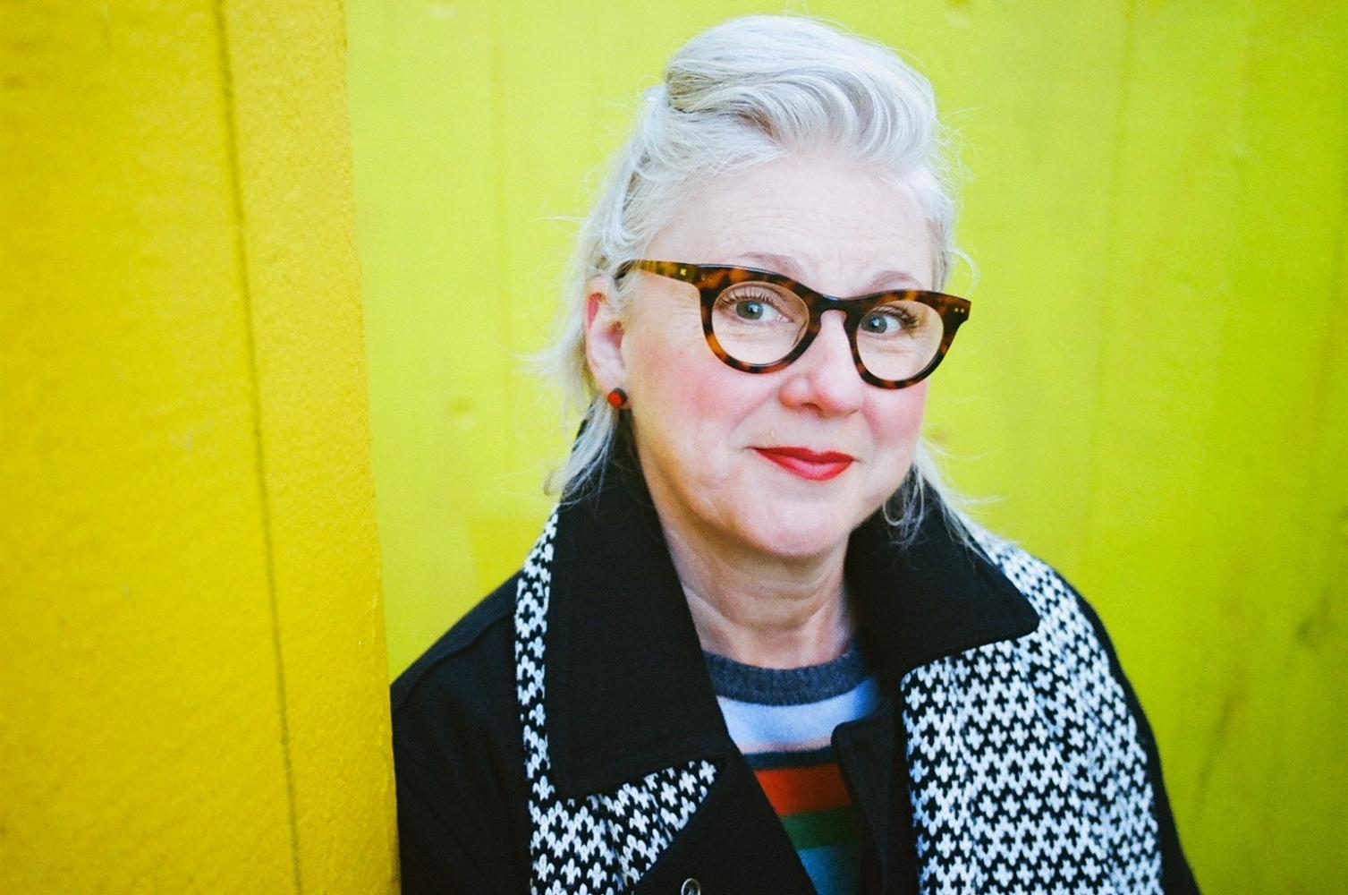 Karen McLeod, Perverse Verse guest, is a women with white hair, black framed glasses standing in front of a yellow background