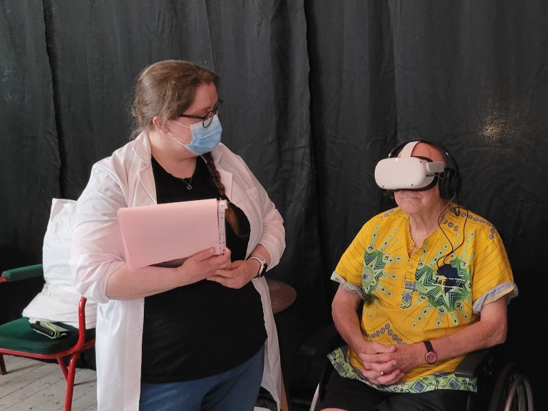 Man in wheelchair wearing yellow top has VR headset and headphones on. Woman in white coat with clipboard watches him.