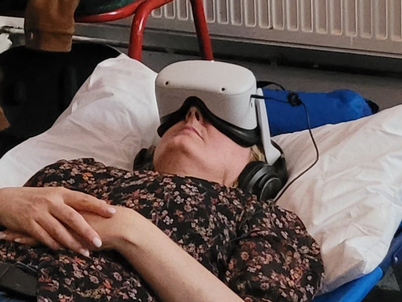 Woman lays on blue surface with head on white pillow.She has VR headset on and headphones. Her arms are crossed, relaxed, across her body.