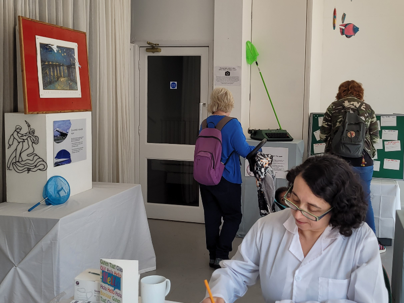 woman seated in white coat in front of area with objects displayed and 2 women looking at them.