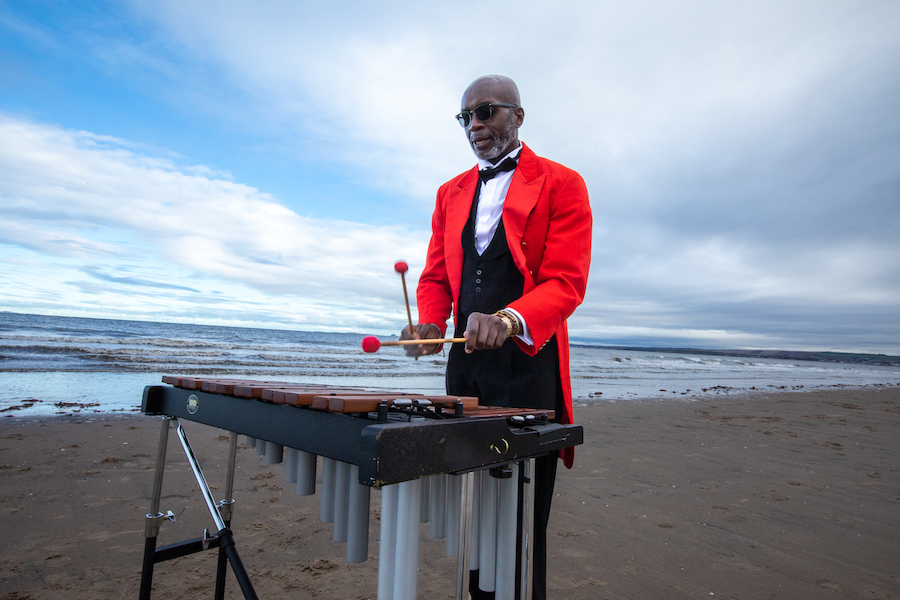 A man in red coat and tails, with a bow tie. He plays a marimba on a beach.