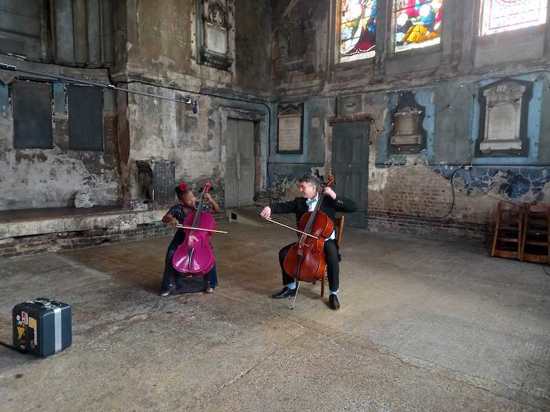 A girl and a man sat next to each other playing cello. They are in an old chapel with distressed, bare brick walls.