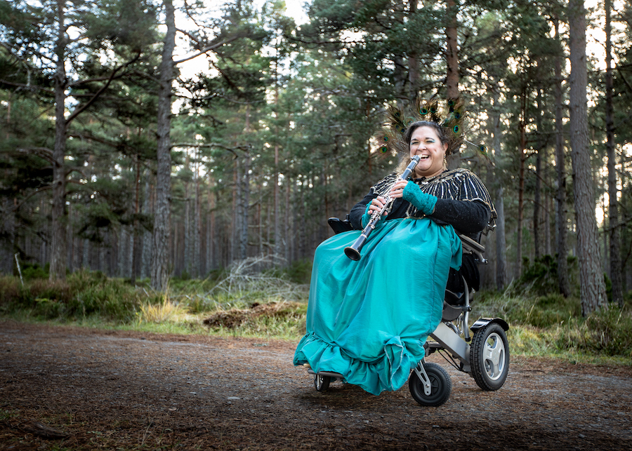 A woman using a wheelchair, outside in a forest. She is laughing and holding a clarinet.