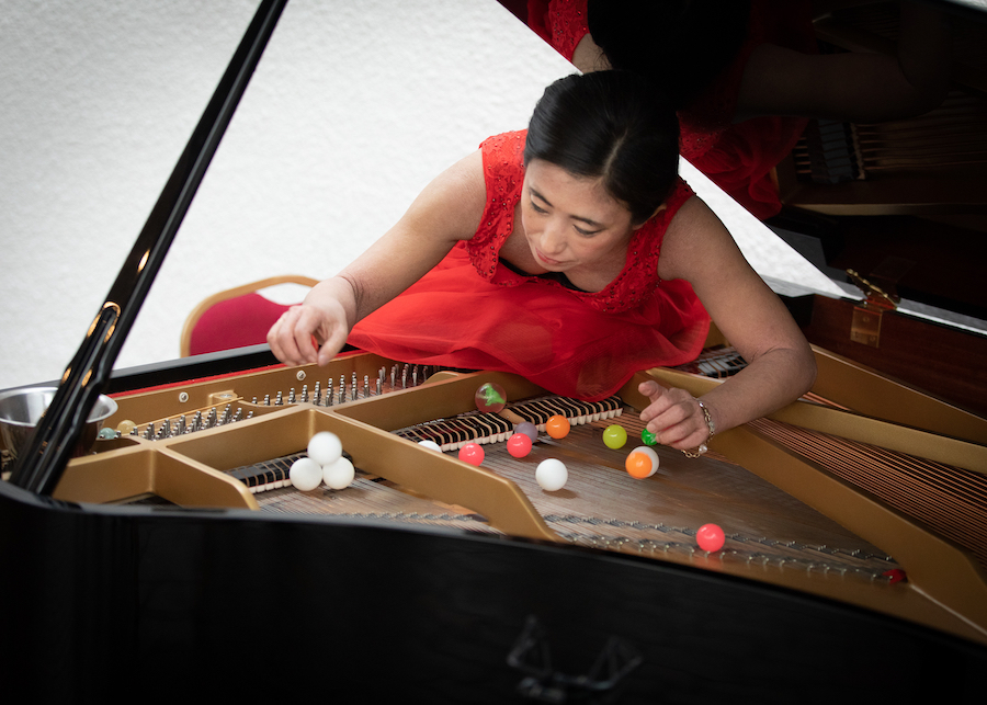 A woman leaning into the top of a piano. She is playing the piano strings with colourful ping pong balls.