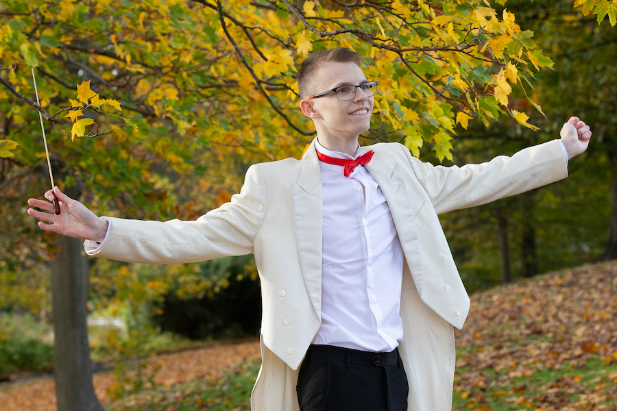 Boy outside, in white coat and tails. His arms are outstretched as he conducts with a baton.