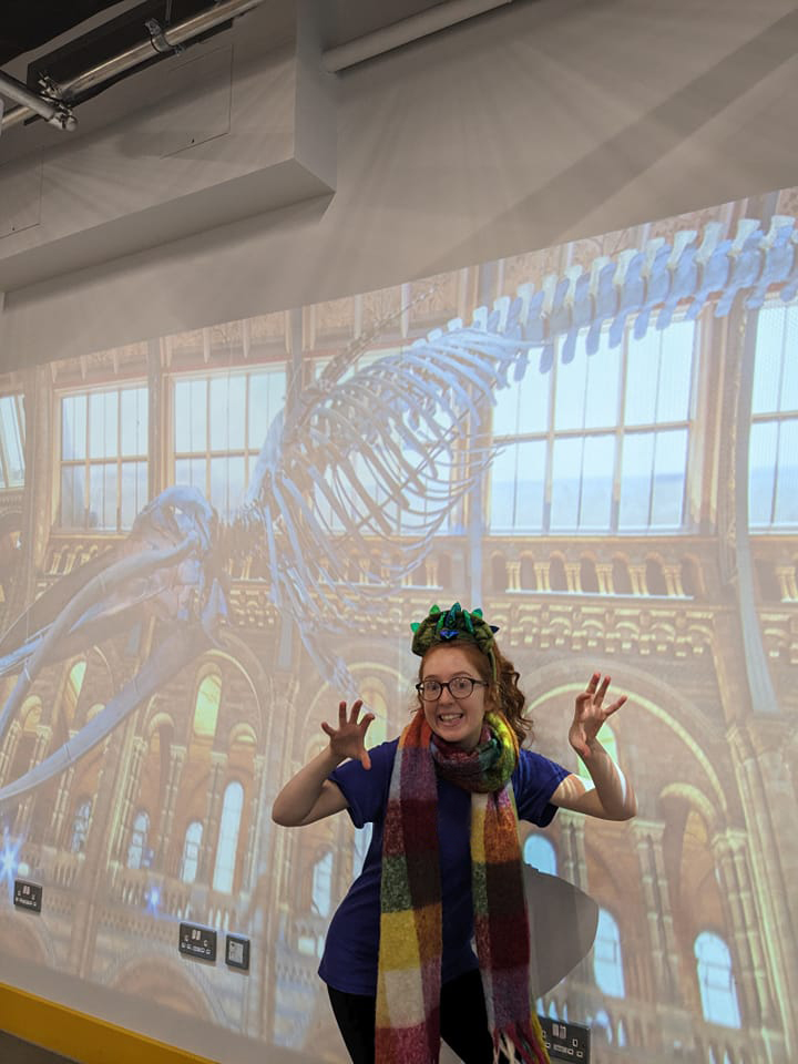 Photo of woman wearing a dinosaur headpiece with large projection of dinosaur bones on wall behind