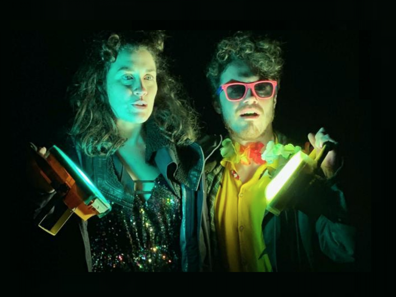 2 figures hold floodlights in their hands. They are dressed in sparkly bright clothing and looking into the distance.