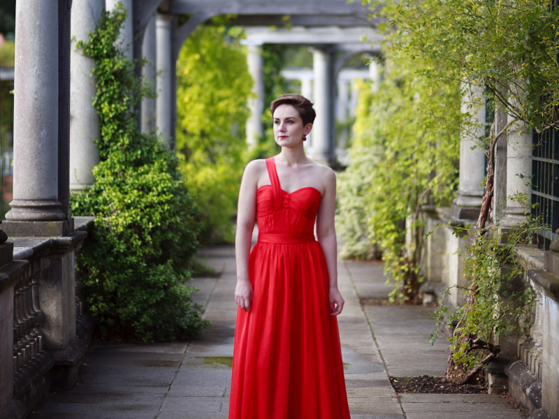 A person in a vibrant red dress stands amidst a serene & beautifully structured garden with stone columns & lush greenery.