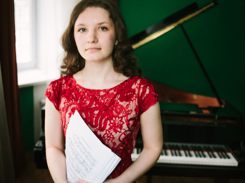 A person in a red lace dress holding sheet music, standing in front of a grand piano by a green wall.