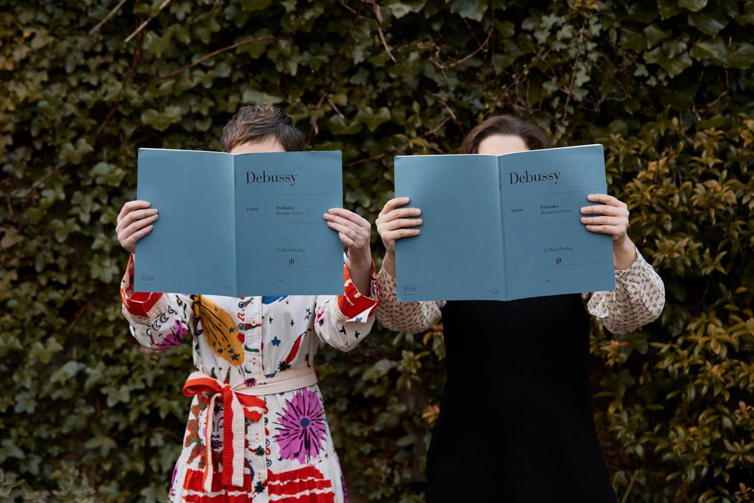 Two women holding blue Debussy music scores in front of their faces