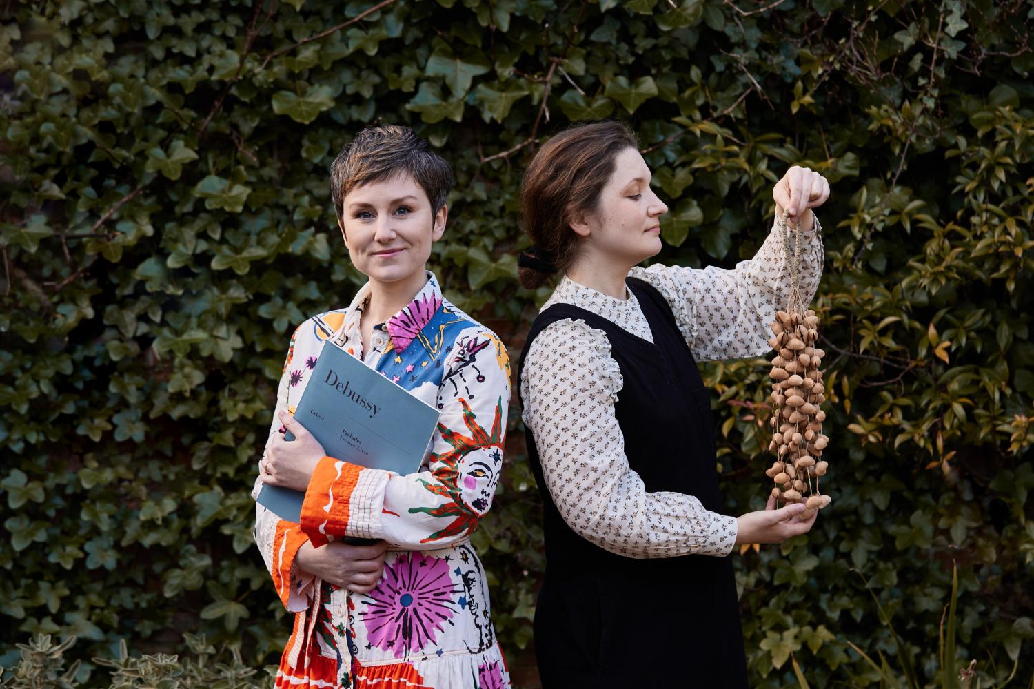 Two women stand side by side. One in portrait holding a seed percussion instrument, the other smiling holding a classical music score.