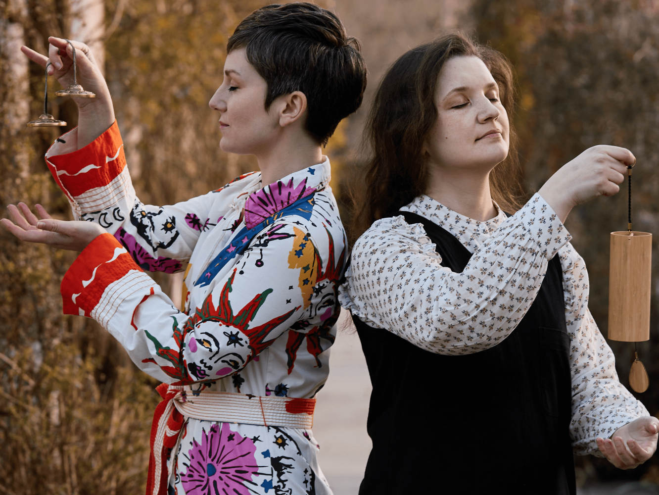 Two women stand back to back with eyes closed in a garden holding different chimes.