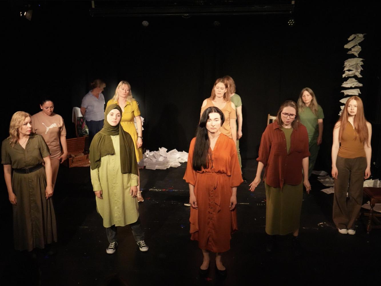 Women stand on stage, they look concerned. They are wearing earth colours and look towards the audience.